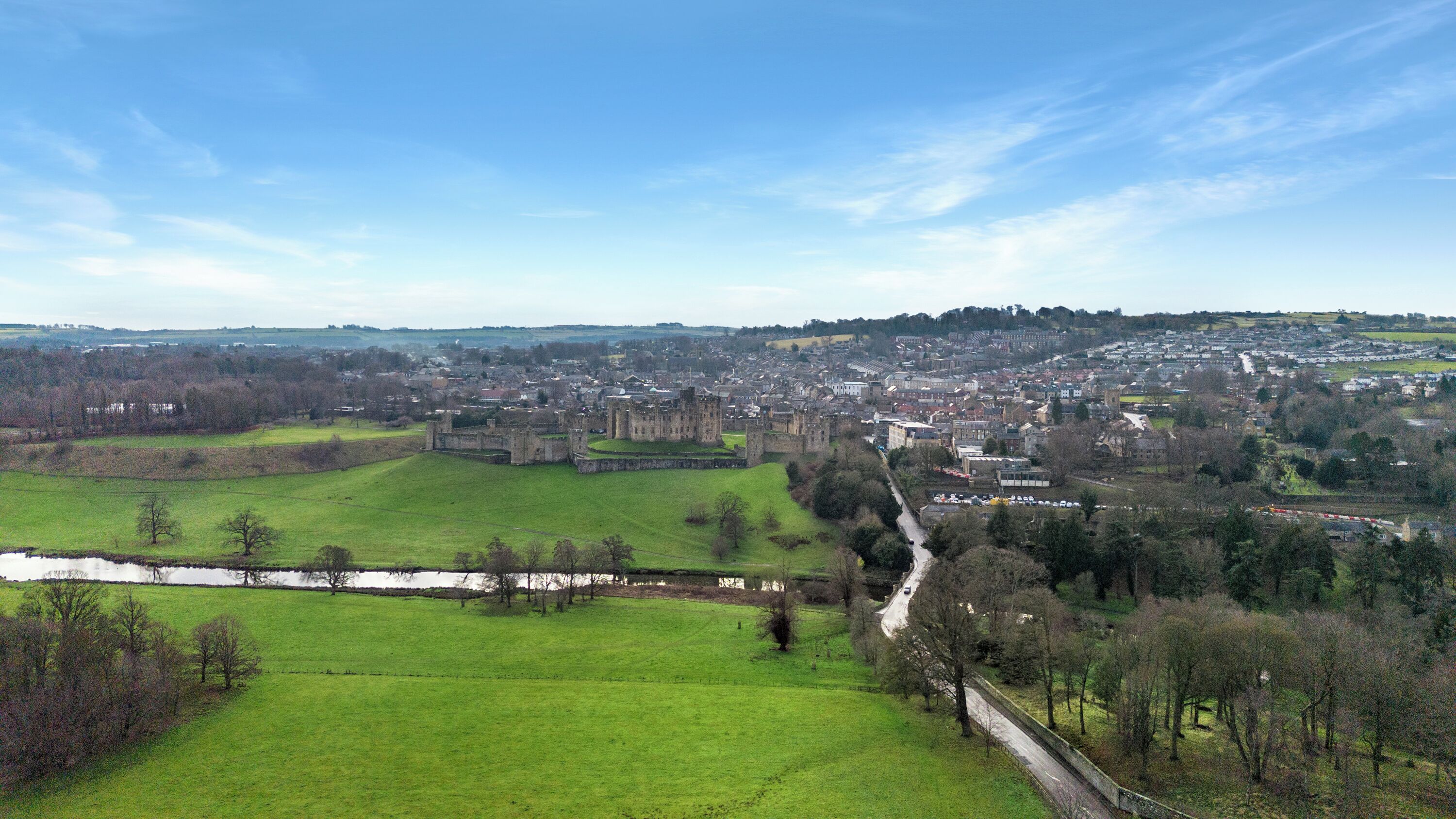 A historic castle surrounded by a town, with green fields and a river in the foreground.