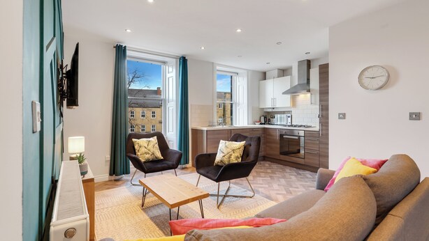 A modern living room with a sofa, two armchairs, a coffee table, and a TV mounted on the wall. There is a kitchen area with cabinets and a stove in the background. - Darjeeling - North East Escapes (Alnwick)
