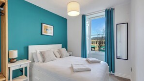 A neatly made bed with white linens and two pillows, a bedside table with a lamp, a framed picture on the wall, and a window with curtains. - Darjeeling - North East Escapes (Alnwick)