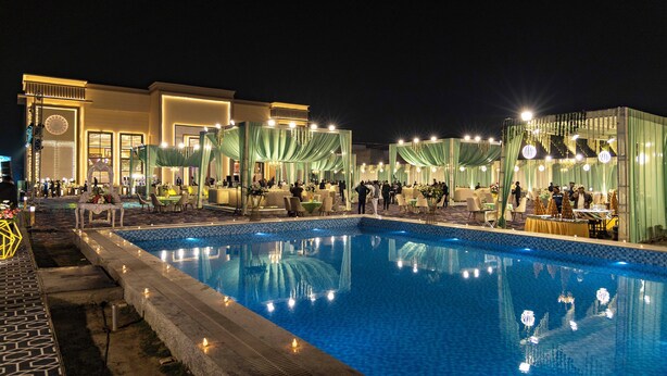 Area piscina illuminata di notte, con vista diretta sulla vasca, zone relax circostanti e un edificio con una finestra circolare.