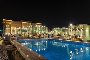 A nighttime pool area with a clear view of the pool, surrounding seating, and a building with a circular window. - THE MADELINNE HOTEL (Muzaffarnagar)