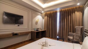 Camera d'albergo con un letto, una TV a parete, una scrivania con una lampada, una sedia e una finestra con tende.