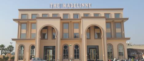 A building with a sign reading "THE MADELINNE" and a parking area with several parked vehicles.