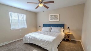 A bedroom with a bed, a ceiling fan, a window with blinds, a bedside table with a lamp, and a wall-mounted artwork.