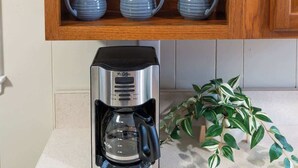 A kitchen with a wooden cabinet containing a green decorative item and three blue mugs. Below the cabinet, there is a coffee maker on a countertop with a potted plant beside it.