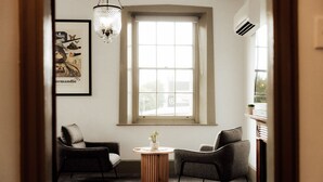 A room with a large window, two armchairs, a small wooden table with a plant, and a chandelier.