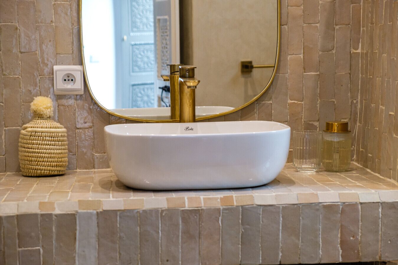 A bathroom with a white vessel sink, gold faucets, a round mirror, and a textured stone wall.