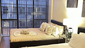 A hotel room with two beds, a large window with blinds, a bedside lamp, and a painting on the wall. - Urban Chic: 2BR + FREE Parking (Birmingham)