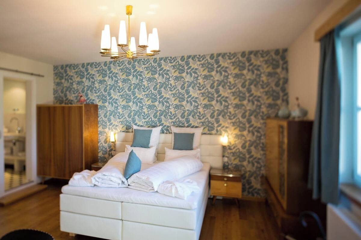 A bedroom with a white bed, blue patterned wallpaper, a chandelier, and wooden furniture.