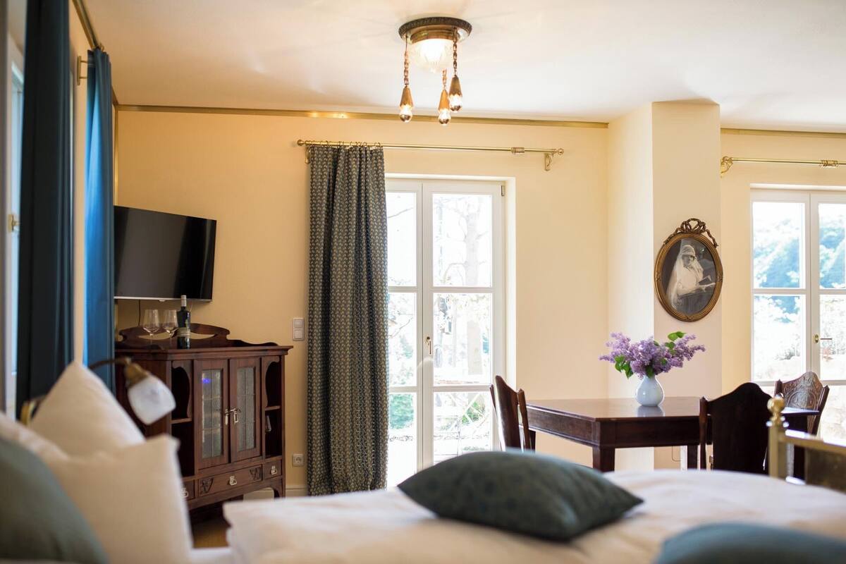 A bedroom with a bed, a TV, a wooden cabinet, a table with a vase, and a window with curtains.