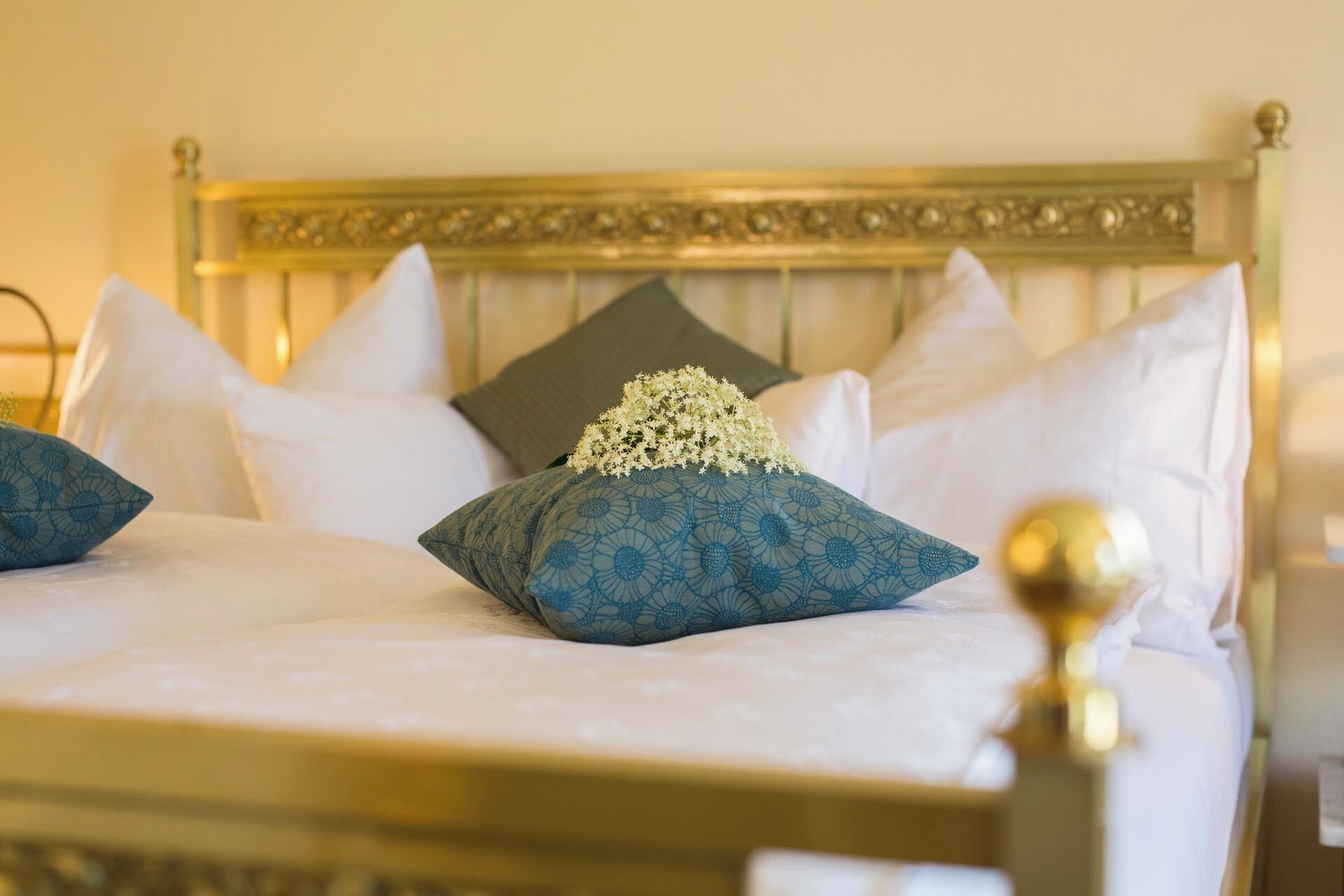 A bed with a gold headboard, white and blue patterned pillows, and a small bouquet of white flowers.