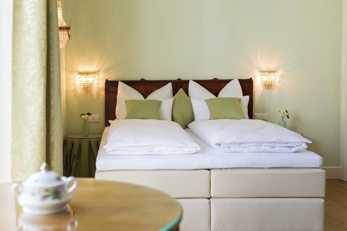 A neatly made bed with white linens and green pillows, a wooden headboard, and two wall-mounted light fixtures.