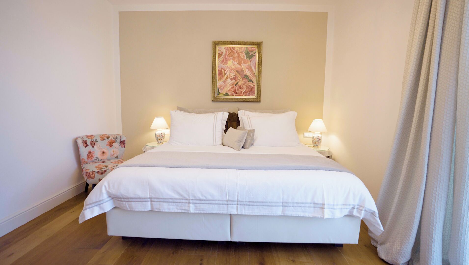 A neatly made bed with white linens, two bedside lamps, a floral armchair, and a painting on the wall.