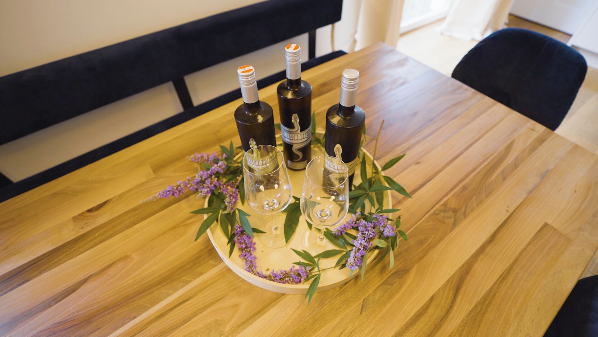 Een houten tafel met drie flessen en twee glaasjes op een dienblad, omringd door paarse bloemen.