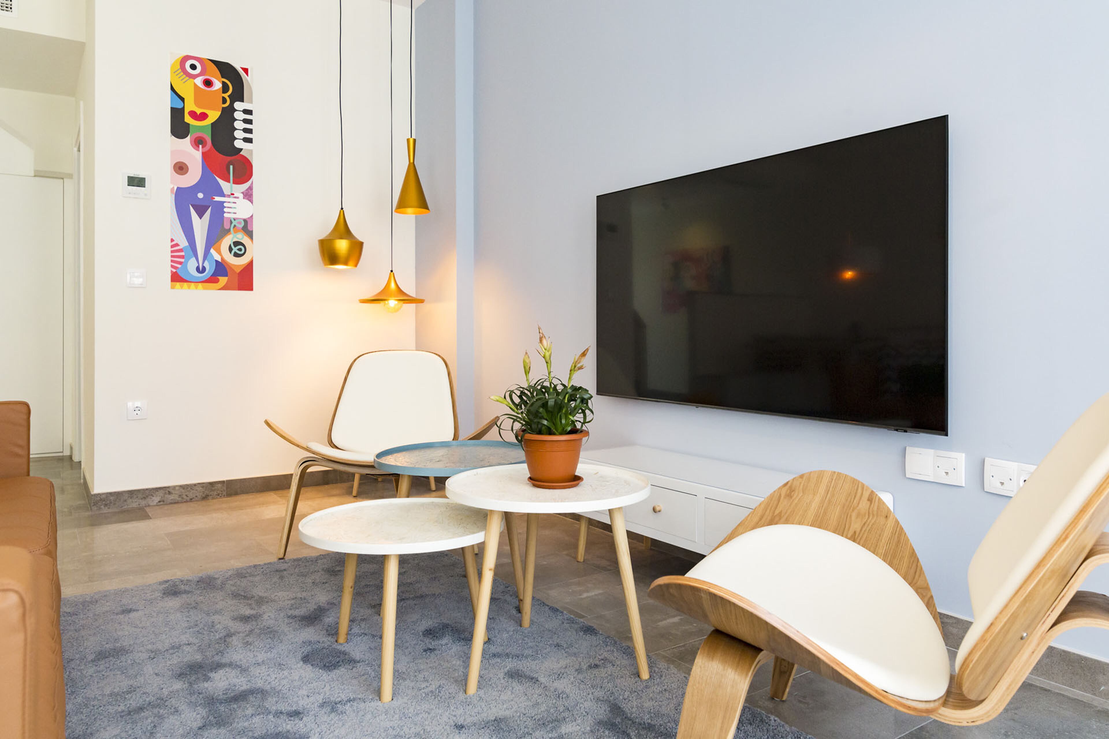 A modern living room with a flat-screen TV, two chairs, a small table with a plant, and a colorful abstract painting on the wall.