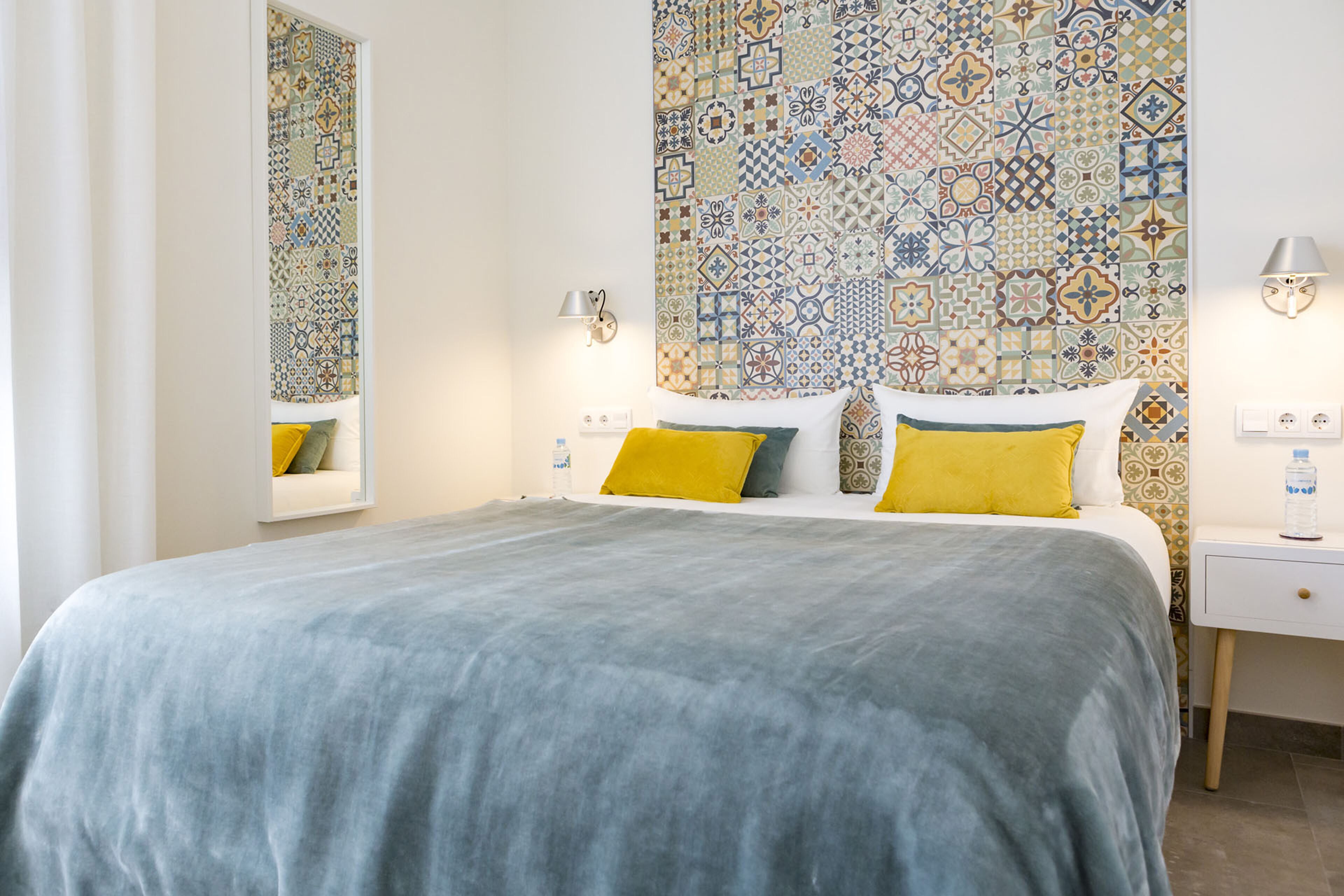 A bedroom with a patterned tile wall, a bed with a grey bedspread, yellow pillows, and a bedside table with a lamp.
