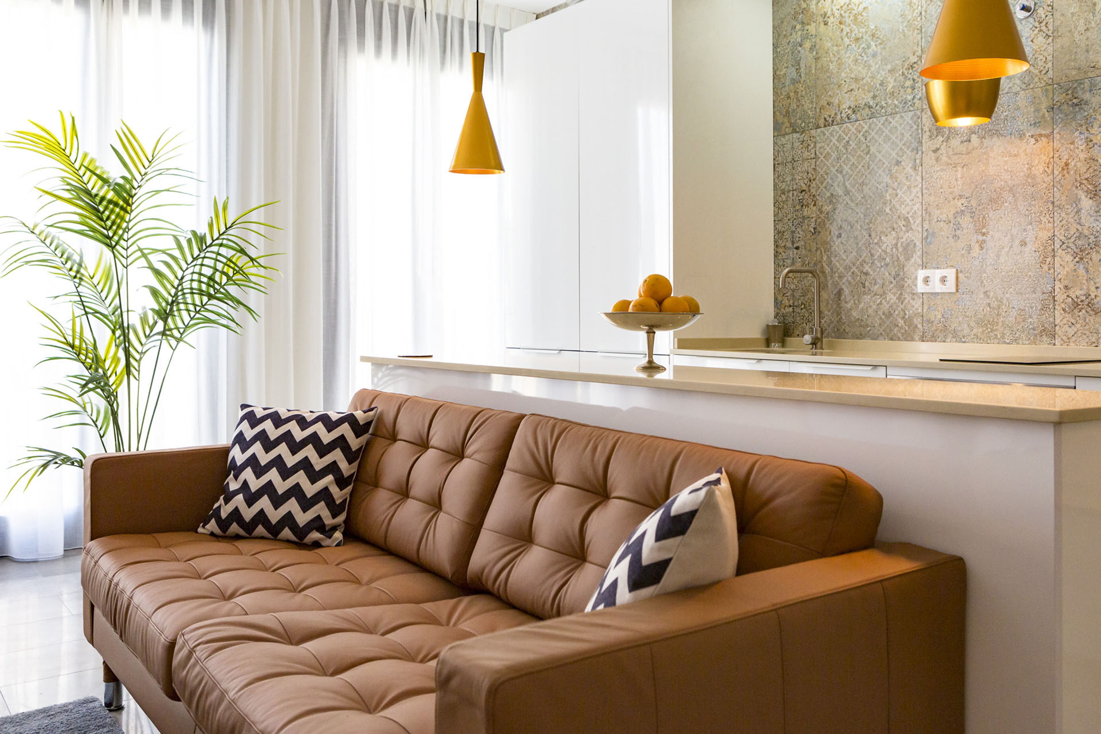 A modern living room with a brown leather sofa, two patterned pillows, a small table with a bowl of fruit, and a potted plant.