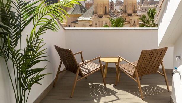 A balcony with two wicker chairs and a small round table, overlooking a cityscape with distinctive architecture. - Solaga - Alta (Málaga)