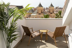 Un balcón con dos sillas de mimbre y una mesita redonda, con vistas a un paisaje urbano de arquitectura singular.