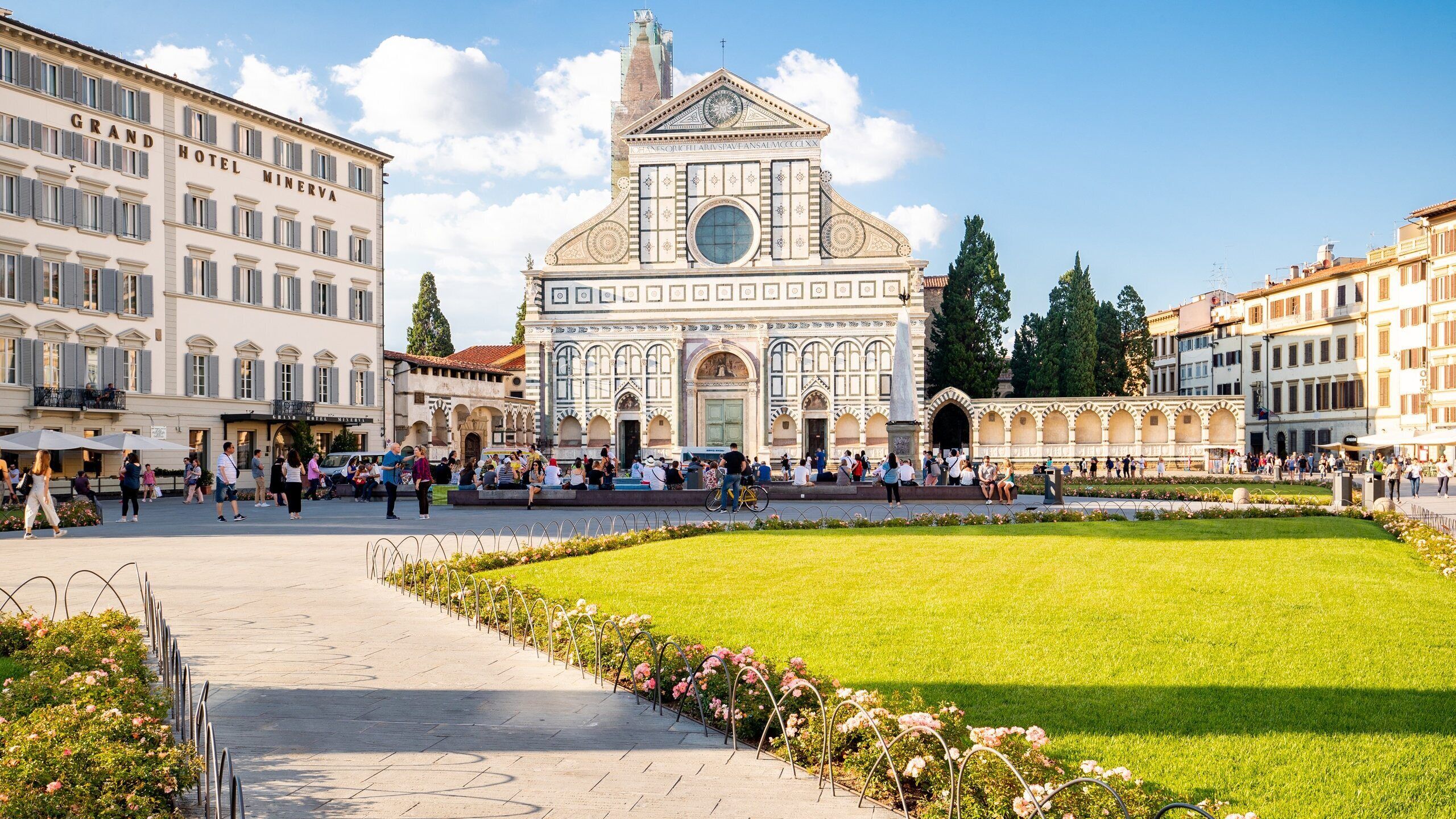 A historic building with a central dome and arched windows, surrounded by a public square with people and greenery.