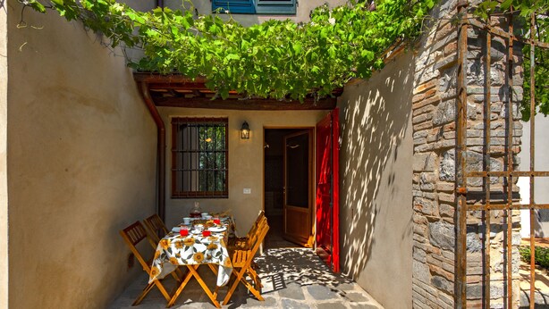 A stone-floored patio with a table and chairs, a window with bars, and a red door.