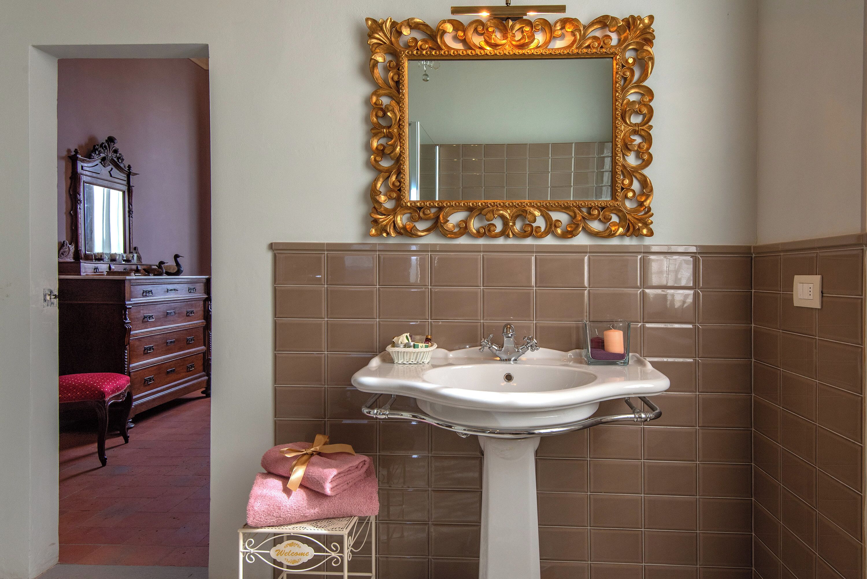 Een badkamer met een wit wastafeltje op voet, een spiegel met gouden lijst en bruine betegelde wanden.