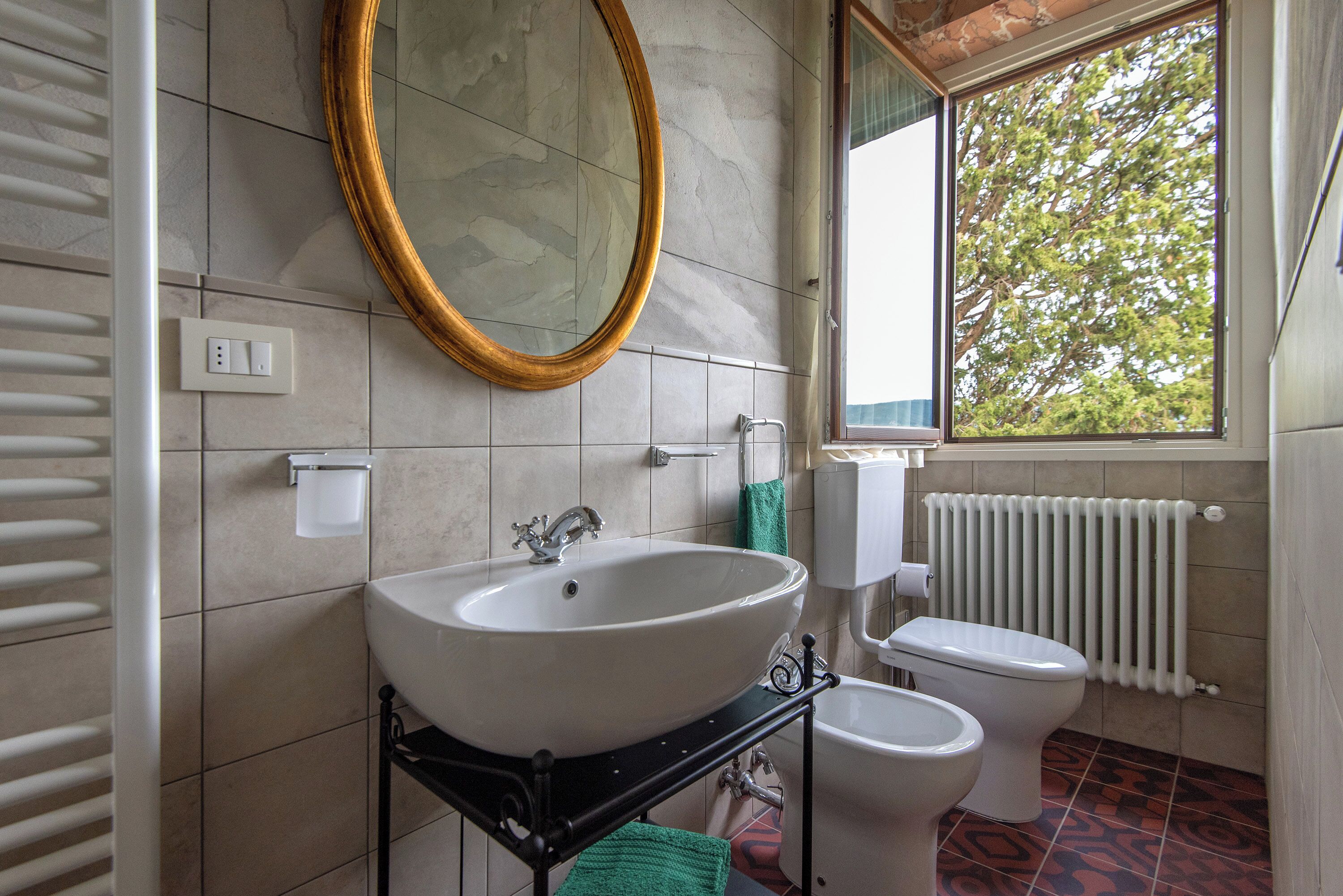 Une salle de bain avec un lavabo blanc, des toilettes, un miroir et une fenêtre donnant sur des arbres.