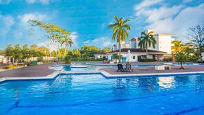 A swimming pool area with lounge chairs, palm trees, and a building with a red roof. - Stylish Oceanfront Condo 3205 | Playa Bejuco + Bright Design + Pool (Bejuco)