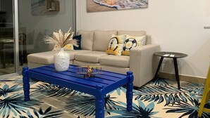 A living room with a sofa, a coffee table, a painting of a beach, and a patterned carpet. - Stylish Oceanfront Condo 3205 | Playa Bejuco + Bright Design + Pool (Bejuco)