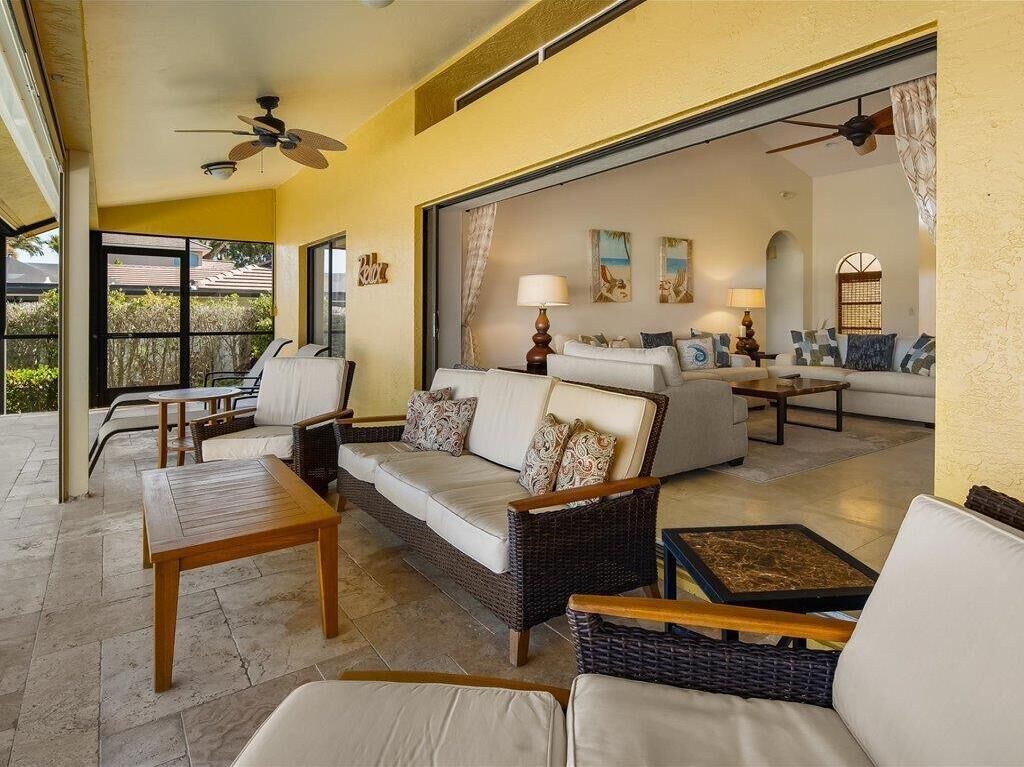 A spacious living area with a sofa set, a coffee table, and a dining table. There is a ceiling fan and a view of the outdoors through large windows.