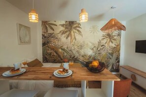 A dining area with a wooden table, two hanging light fixtures, and a tropical mural on the wall. - Studio Ti Zero (Entre-Deux)