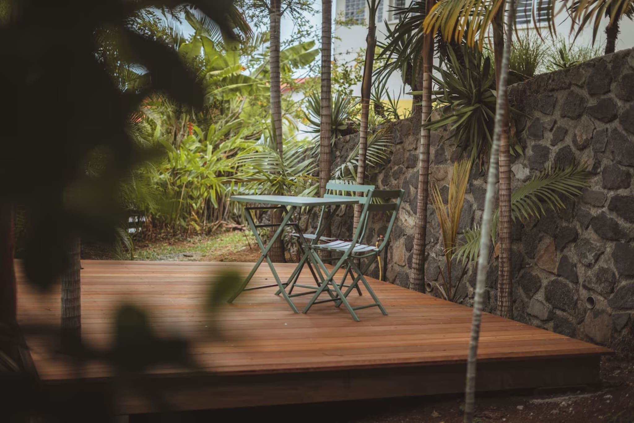 A wooden deck with a green metal table and chairs, surrounded by tropical plants and a stone wall.