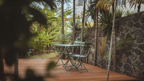 A wooden deck with a green metal table and chairs, surrounded by tropical plants and a stone wall. - Studio Ti Zero (Entre-Deux)
