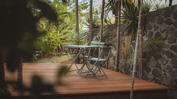 A wooden deck with a green metal table and chairs, surrounded by tropical plants and a stone wall.