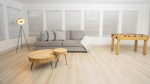 Ein Wohnzimmer mit grauem Sofa, zwei Holztischen, einem Tischfußballtisch, einer stehenden Stehlampe und einem Deckenventilator.