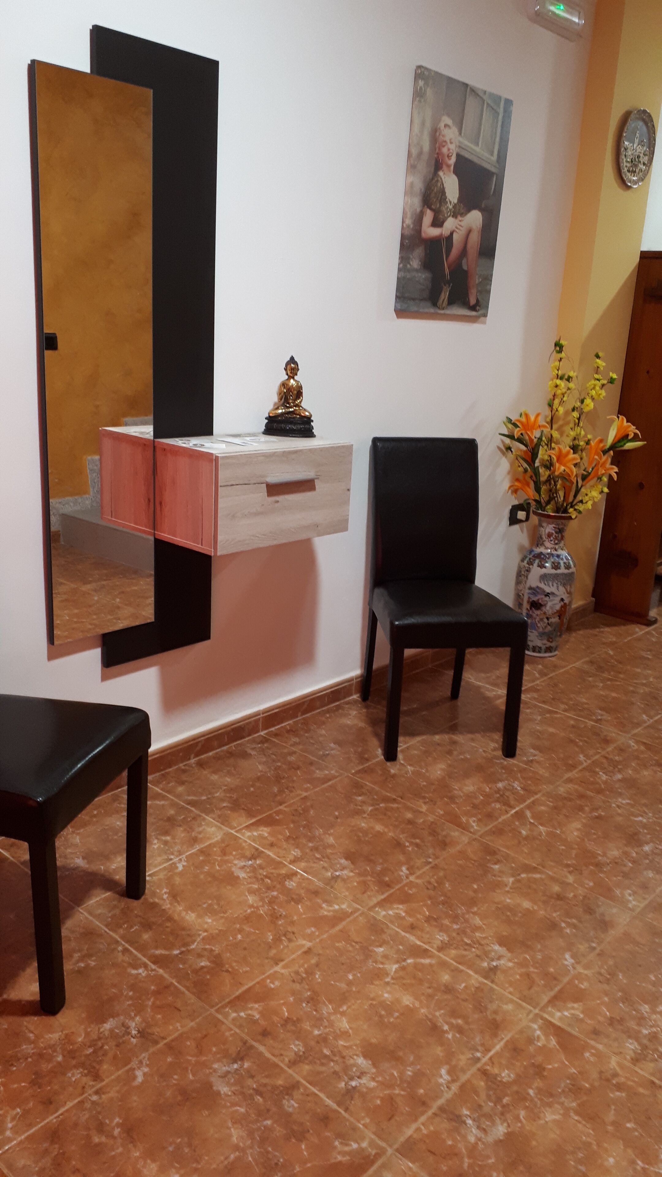 A room with a wooden cabinet, a black chair, a mirror, a Buddha statue, a framed picture, and a vase with flowers.