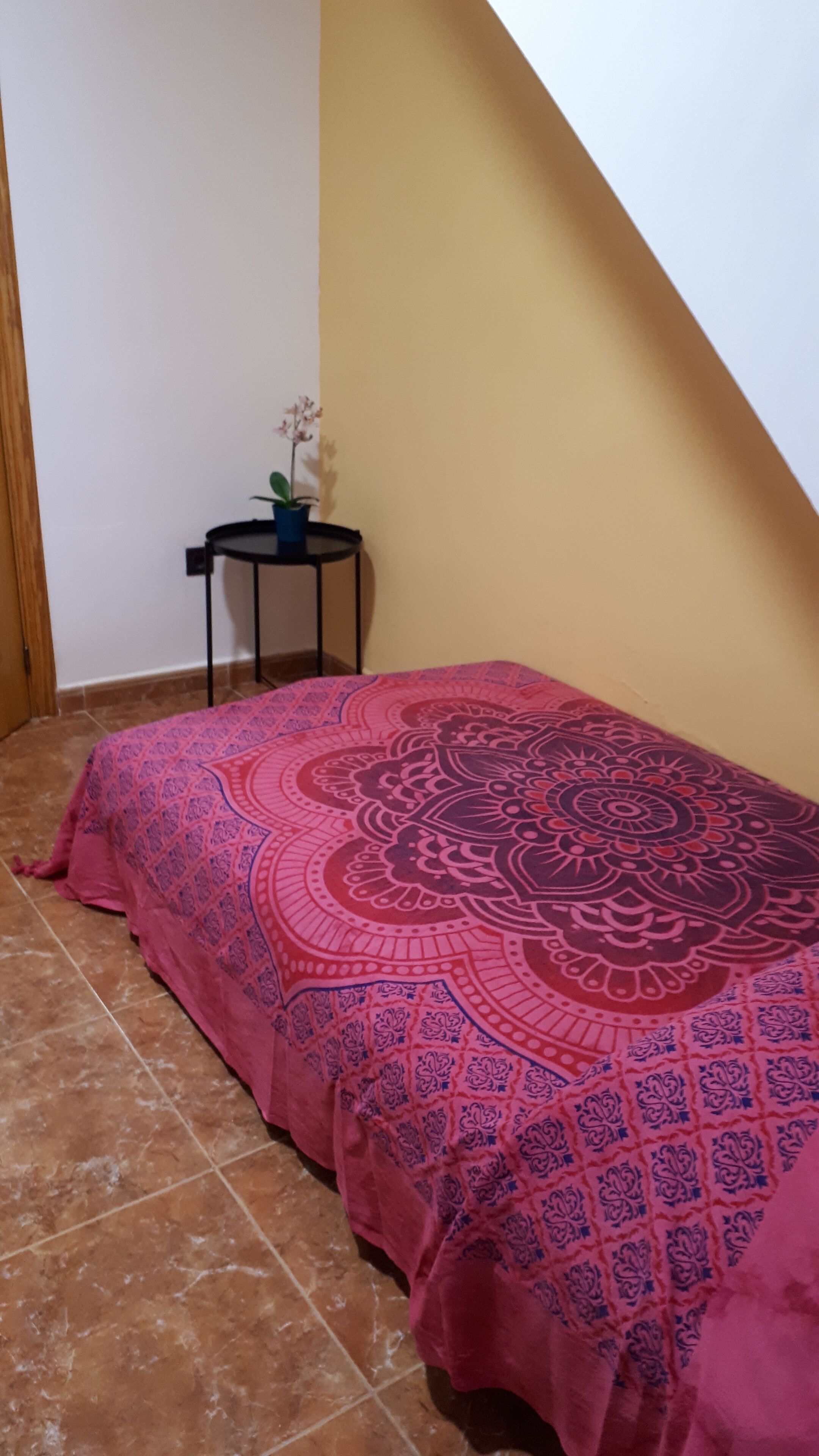 A room with a bed covered in a pink patterned bedspread, a small round table with a potted plant, and a tiled floor.