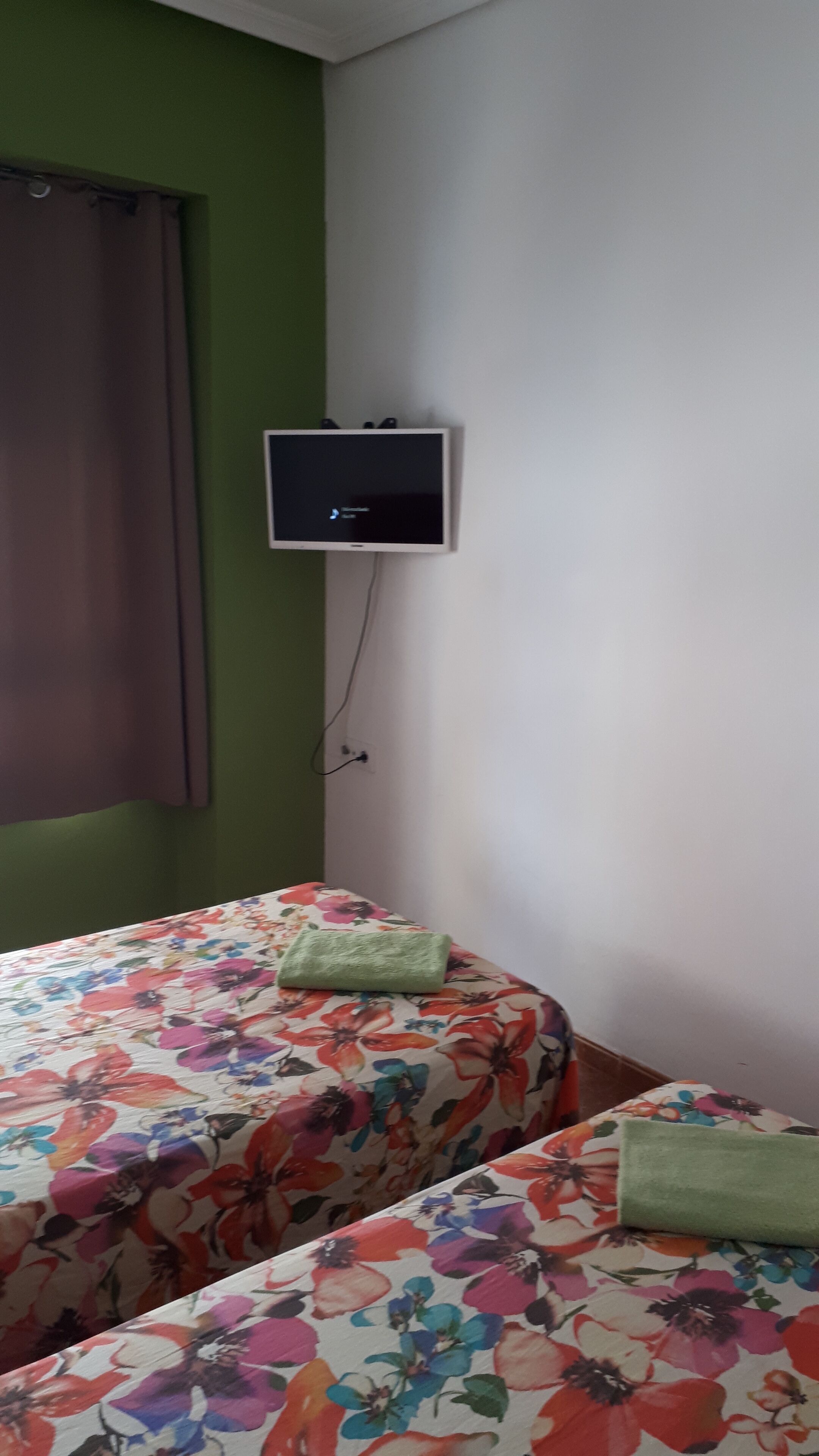 Une chambre avec deux lits, chacun recouvert d’une couvre-lit à motifs floraux, un téléviseur fixé au mur et une fenêtre avec un rideau marron.