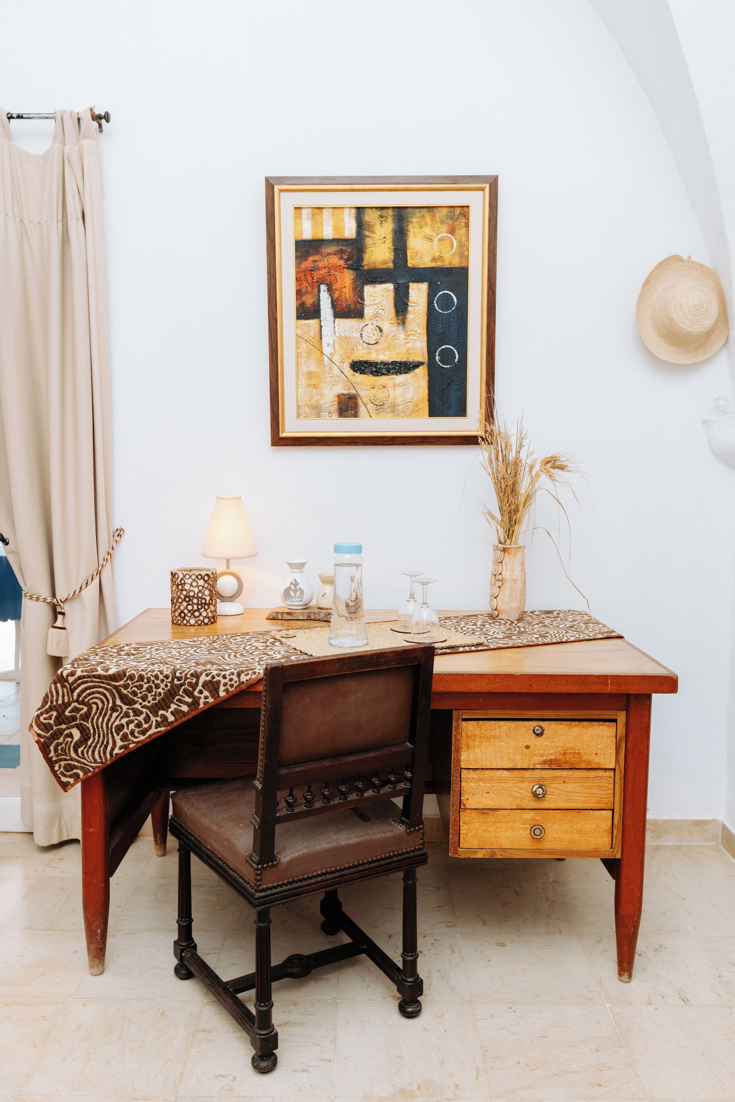 Una scrivania in legno con una tovaglia decorata, una sedia, una lampada, un vaso con piante essiccate e un quadro astratto incorniciato appeso al muro.