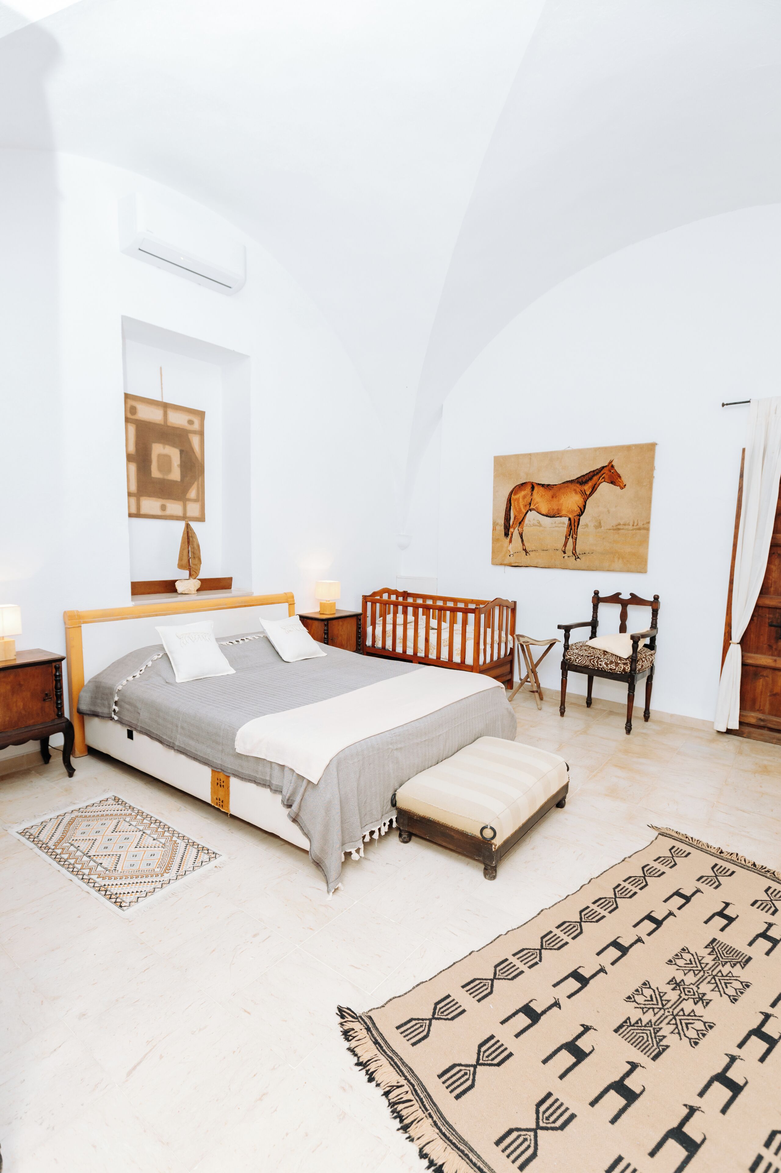 A bedroom with a bed, a chair, a rug, a painting, and a small table with a lamp.