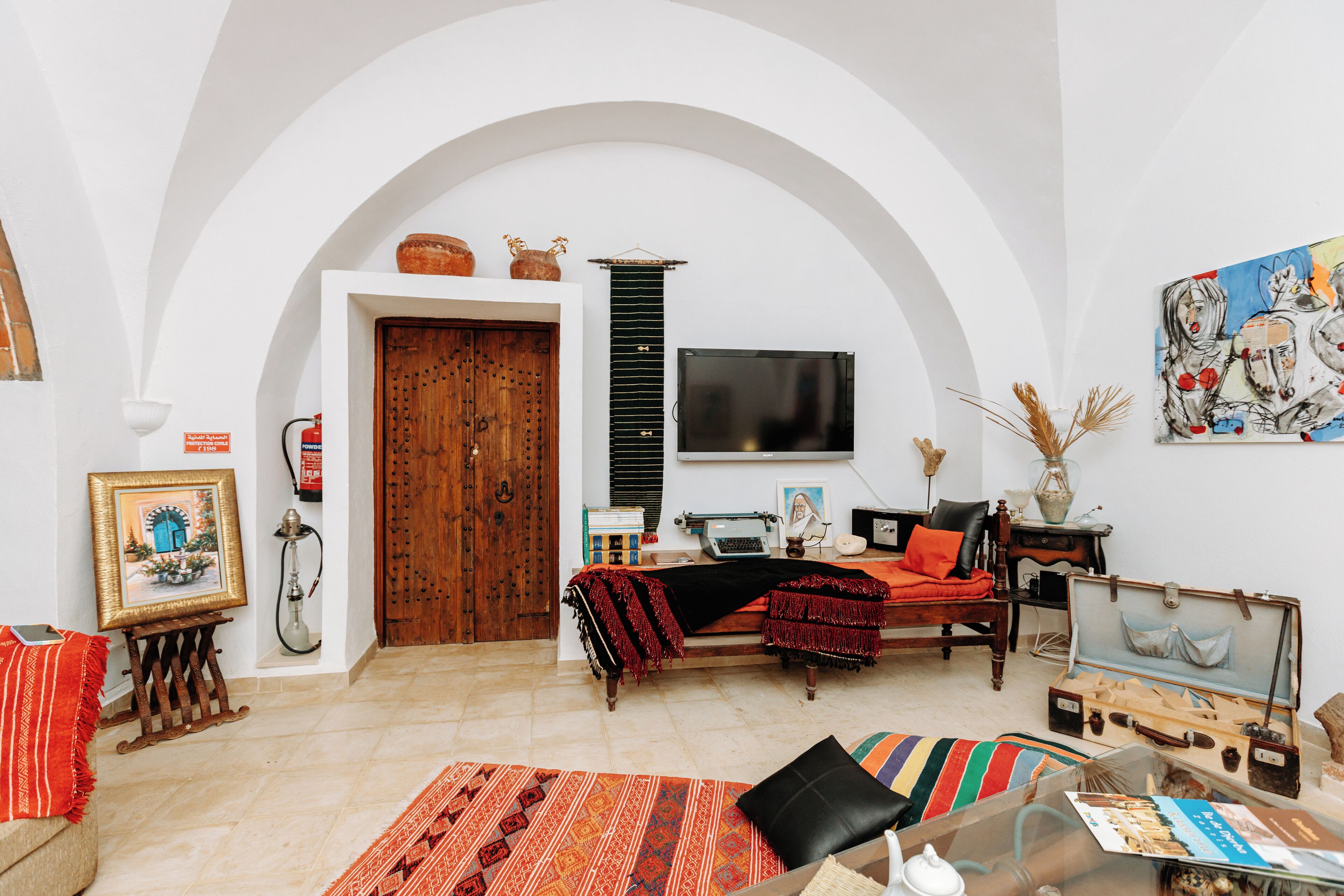 Un salón con sofá, una mesa de centro y un televisor. Se observa una puerta de madera y una pared blanca con un cuadro.