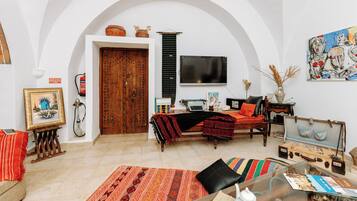 Un salón con sofá, una mesa de centro y un televisor. Se observa una puerta de madera y una pared blanca con un cuadro.