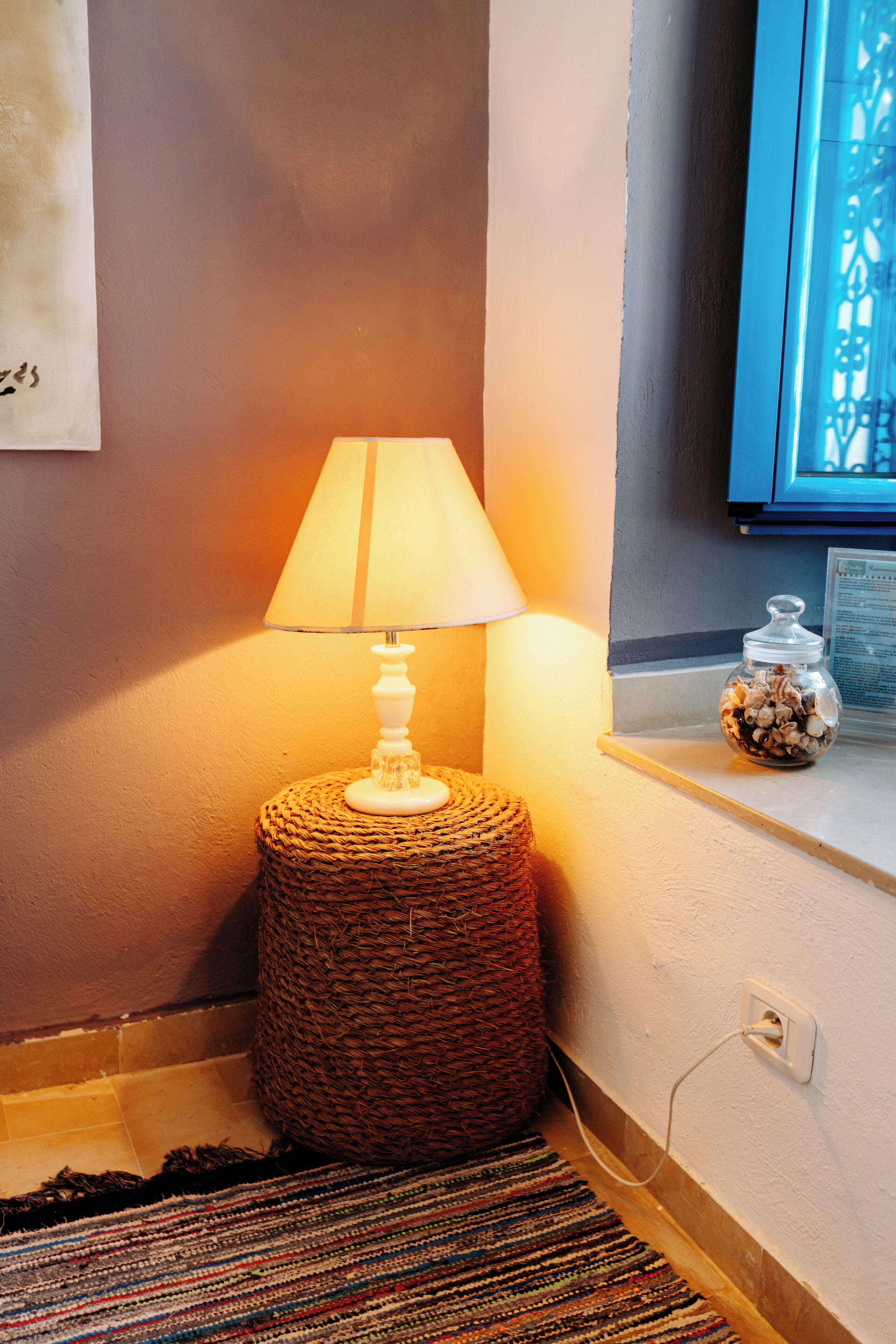 A lamp on a woven stool, a textured rug, and a small jar on a ledge.