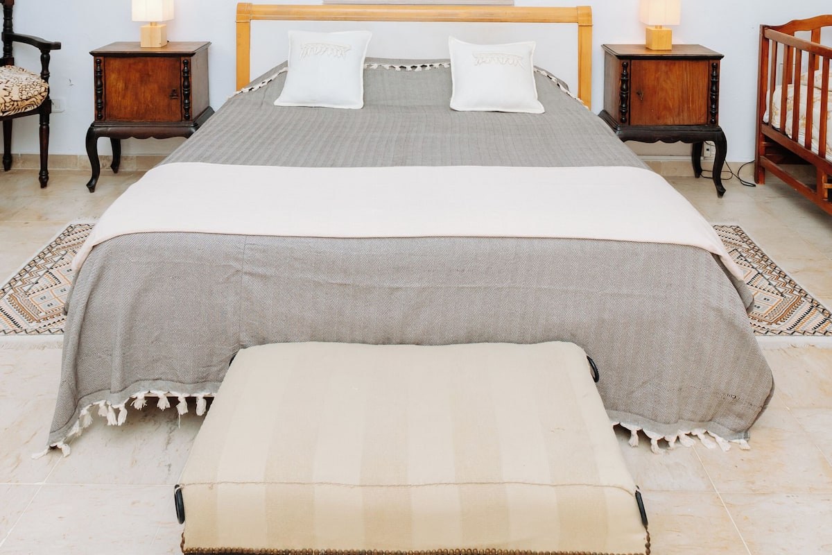 Une chambre à coucher bien aménagée, avec un grand lit, deux tables de chevet avec des lampes, une tête de lit en bois, un climatiseur fixé au mur et un tableau encadré au-dessus du lit.