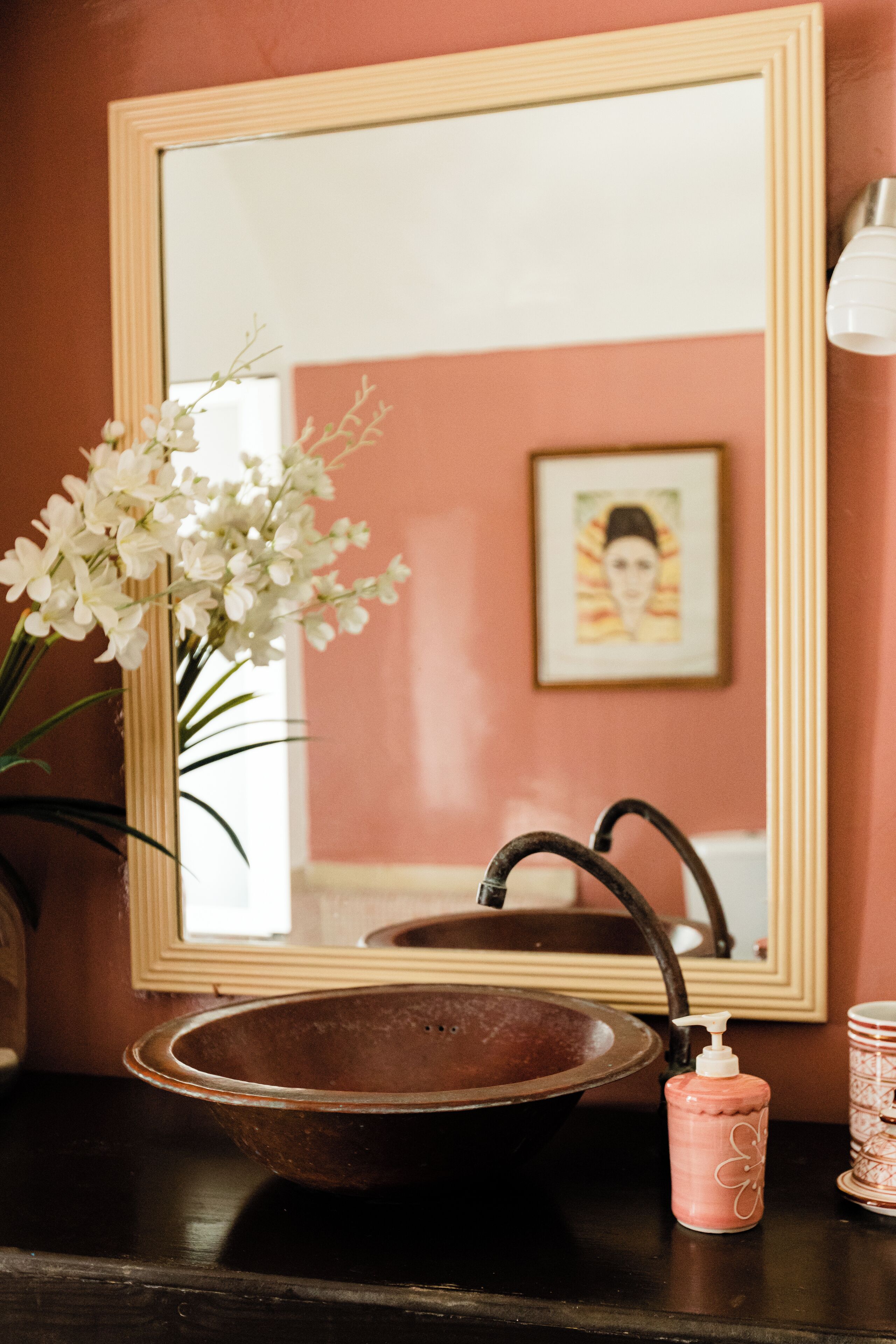 Un baño con lavamanos de madera, un espejo, un cuadro en la pared y un florero con flores blancas.