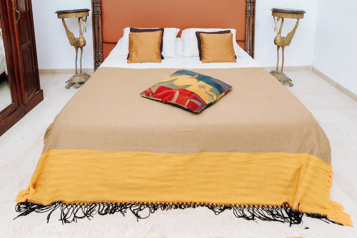 Une chambre à coucher avec une tête de lit en bois, un lit recouvert d’une couverture beige et agrémenté d’oreillers, un plaid coloré et deux tables de chevet avec des lampes.