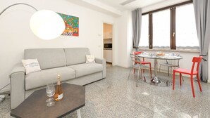 En moderne stue med en grå sofa, et sofabord med en flaske og glas, et spisebord med røde stole, og et stort vindue med gardiner.