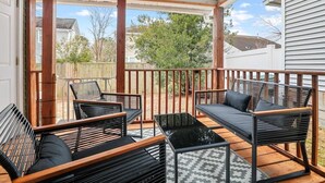 Un patio techado con suelo de madera, muebles negros y de madera, y vistas a un barrio residencial.