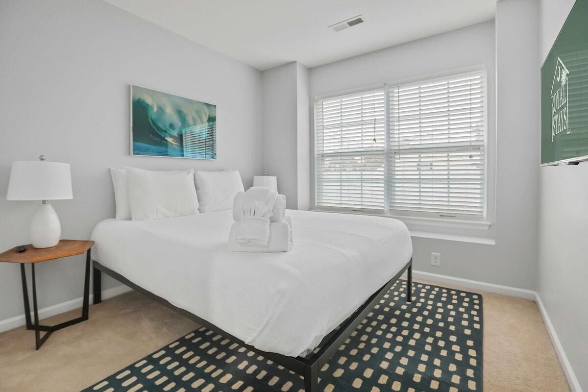 A hotel room with a large bed, a bedside table with a lamp, a window with blinds, and a painting on the wall.