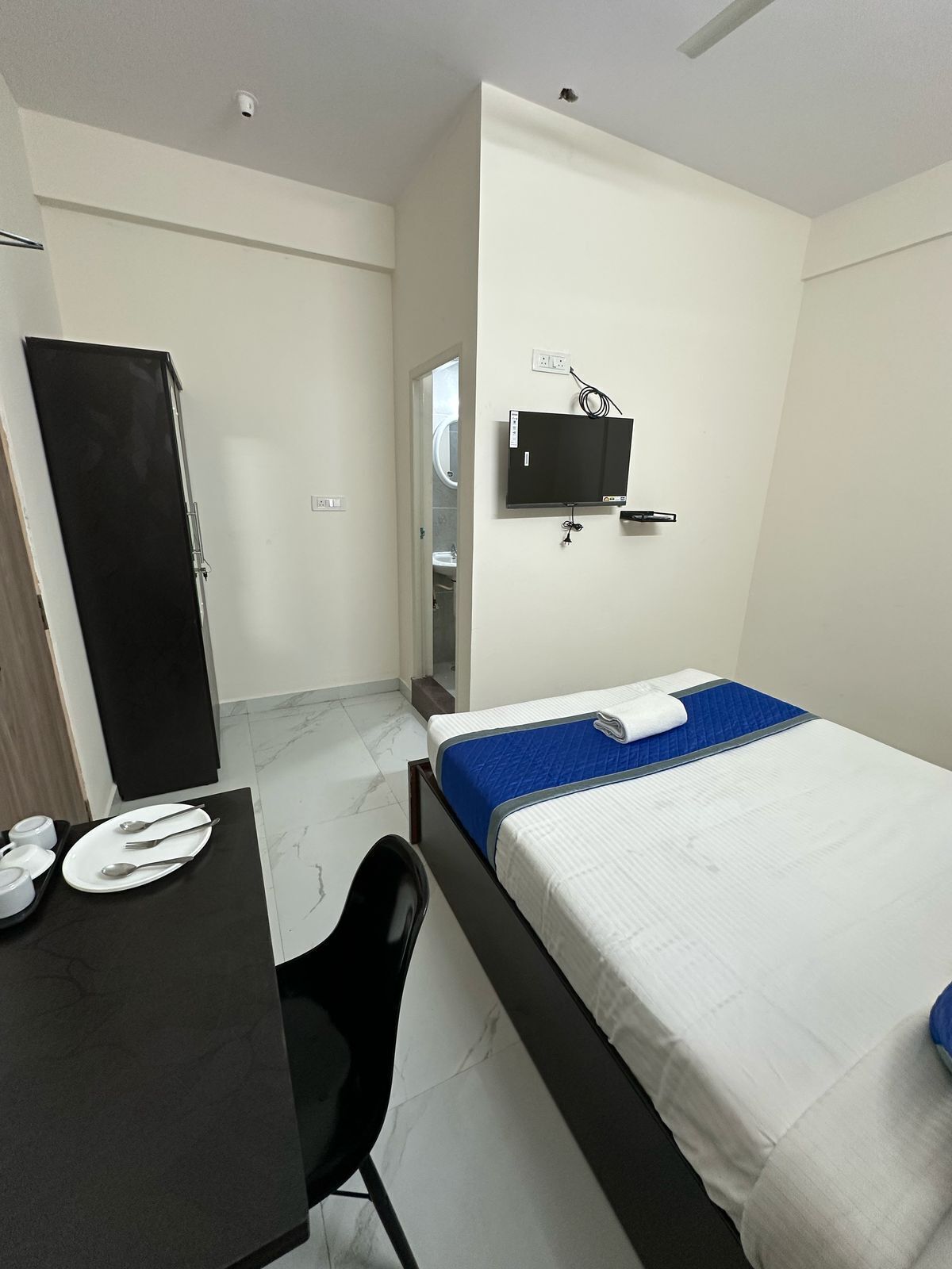 Una habitación de hotel con cama, escritorio, silla, televisor montado en la pared y puerta que da a un baño.